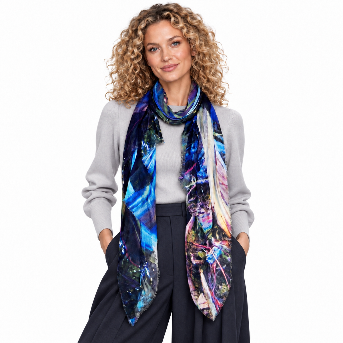 A woman with curly blonde hair models a vibrant, multicolored abstract print scarf. She is wearing a light gray long-sleeve top and dark navy wide-leg trousers, standing against a solid white studio background.