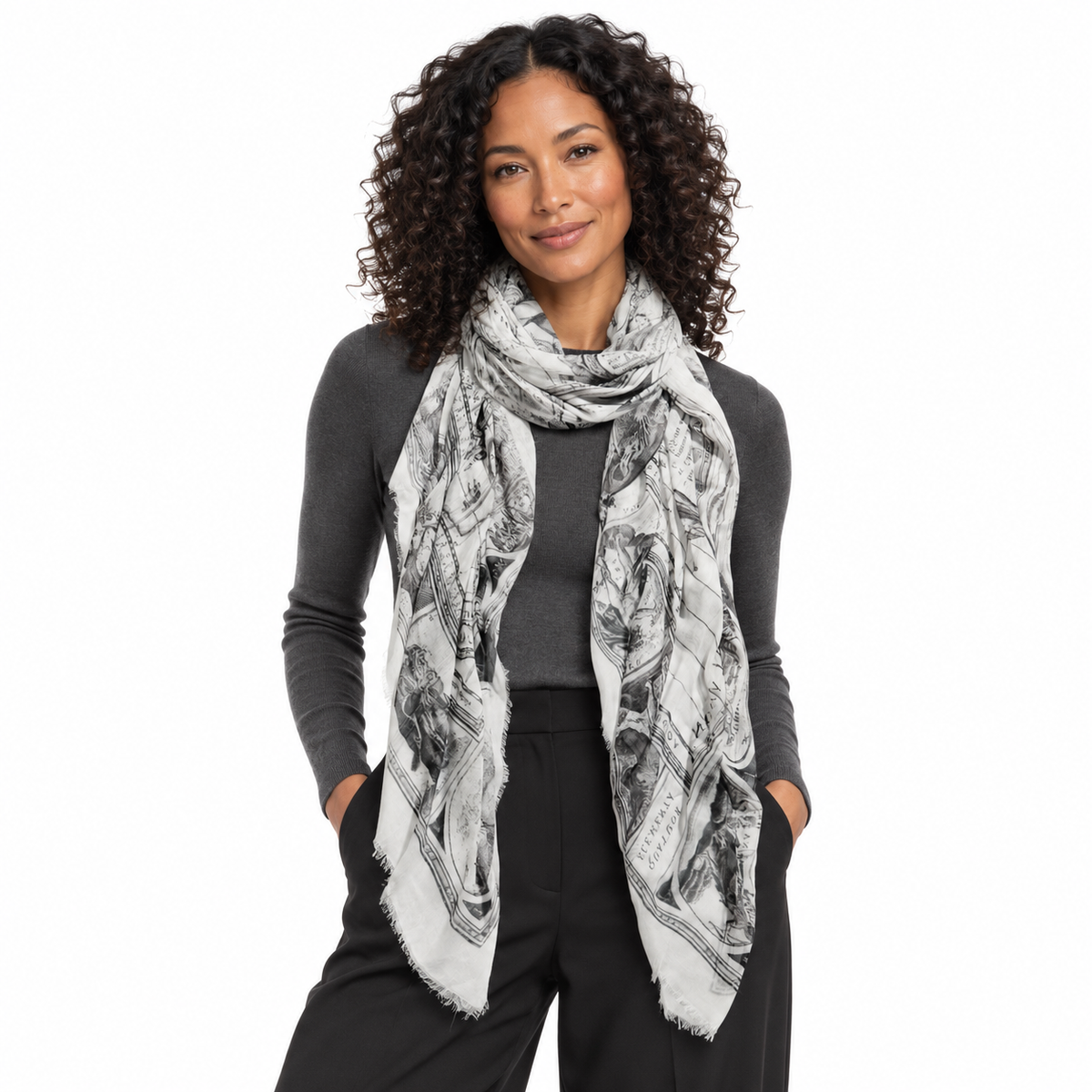 A woman with curly dark hair models a light gray and black printed scarf over a dark gray long-sleeve top and black pants against a plain white background.