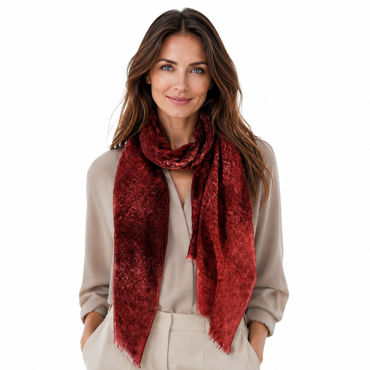 A woman with long brown hair models a textured, deep red scarf wrapped around her neck, paired with a beige top and white trousers, against a solid white background.